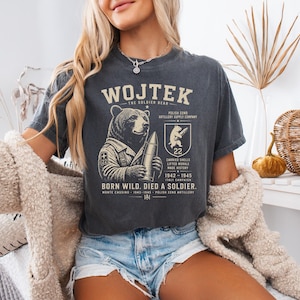 May include: A charcoal gray short-sleeve t-shirt featuring a graphic of a bear holding an artillery shell. The text reads WOJTEK, THE SOLDIER BEAR, POLISH 22ND ARTILLERY SUPPLY COMPANY, 22, CARRIED SHELLS LIFTED MORALE MADE HISTORY, 1942-1945, BORN WILD. DIED A SOLDIER.