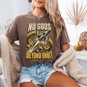 May include: A brown short-sleeve t-shirt featuring a graphic print of a rocket ship breaking through a yellow ring. The text "NO GODS" appears above the rocket, and "BEYOND ORBIT" is printed below in a bold, distressed yellow font.