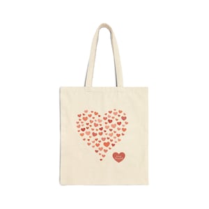 Puede incluir: Bolso tote de color crema con un gran corazón hecho de corazones rojos más pequeños. Un corazón rojo más pequeño con las palabras "Love Yourself" está debajo del corazón más grande. El bolso tiene dos asas.