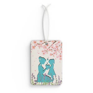 Puede incluir: Un ambientador de coche blanco con una silueta azul de una madre y una hija tomadas de la mano bajo un cerezo en flor rosa. El diseño está recortado de la tarjeta, creando un efecto de capas.