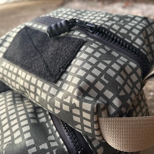 May include: Close-up of two square bags with a repeating grid pattern in shades of gray and olive green. Each bag has a black zipper, a black velcro patch, and a tan strap. The bags are made of a durable, textured fabric.