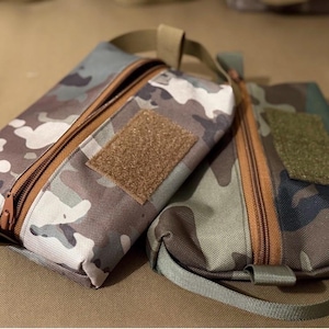 May include: Two camouflage-patterned pouches with brown zippers and Velcro patches. One pouch is rectangular, the other is more compact. Both have a loop handle. The pouches are in shades of brown, green, and white.
