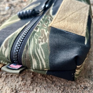 May include: A close-up shot of a camouflage pouch with a black zip and a tan velcro patch. The pouch has a tiger stripe pattern in green, brown, and black. An American flag tag is visible.