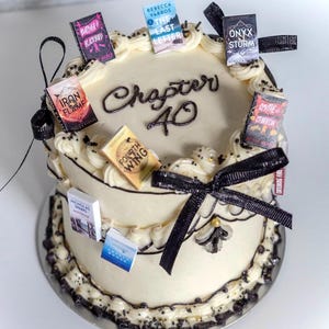 May include: A round, two-tiered cake with white frosting, decorated with black accents and miniature book toppers. The cake is inscribed with "Chapter 40" in elegant script and adorned with black ribbon.