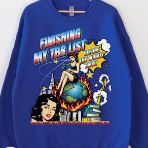 Puede incluir: Sudadera azul real con un gráfico de arte pop. El gráfico presenta el texto "FINISHING MY TBR LIST BEFORE THE WORLD ENDS!" encima de una mujer leyendo en un globo terráqueo rodeado de llamas, con un paisaje urbano y el rostro de una mujer a la izquierda.