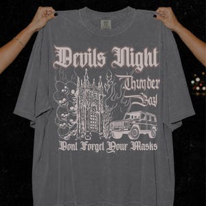 May include: A charcoal gray short-sleeve t-shirt featuring a graphic print with the text Devils Night, Thunder Bay, and Dont Forget Your Masks. The design includes a stack of skulls, a gothic building, flames, and an off-road vehicle.