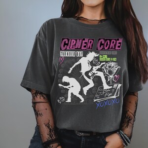 May include: A dark gray t-shirt with a graphic print of two figures playing guitars. The text "CIRCUS CORE" is printed in pink and white. The text "THUNDER BAY" is printed in white. The text "EVENT LIFE ENDS" is printed in white. The text "12+ ONLY" is printed in white. The text "DOORS OPEN 6 PM" is printed in white. The text "From Mika To Ryan" is printed in blue and white. The text "XOXOXO" is printed in white.