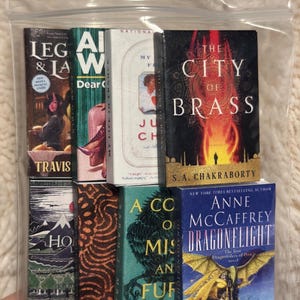 Peut inclure: Une collection de livres miniatures, dont des titres comme "The City of Brass" et "Dragonflight", est pr&eacute;sent&eacute;e dans un sac en plastique transparent. Les livres pr&eacute;sentent des couvertures color&eacute;es avec divers motifs et textes, repr&eacute;sentant diff&eacute;rents genres.