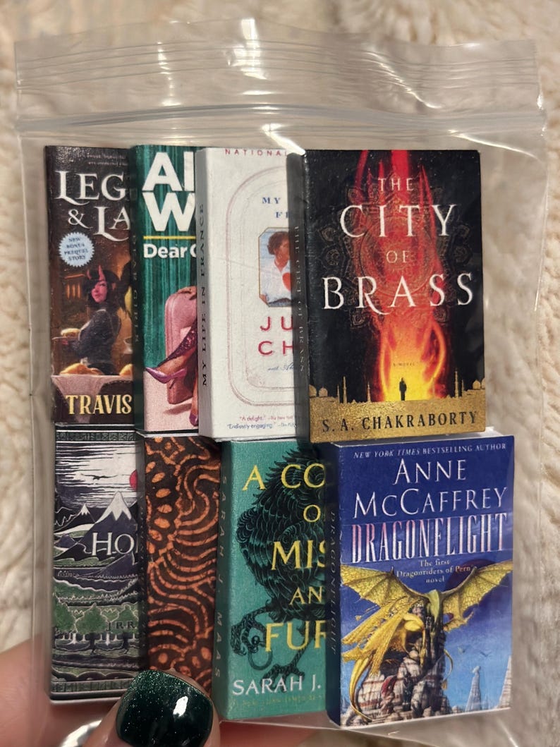 Peut inclure: Une collection de livres miniatures, dont des titres comme "The City of Brass" et "Dragonflight", est pr&eacute;sent&eacute;e dans un sac en plastique transparent. Les livres pr&eacute;sentent des couvertures color&eacute;es avec divers motifs et textes, repr&eacute;sentant diff&eacute;rents genres.