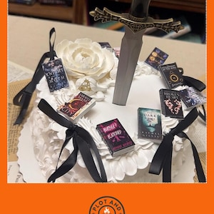 Peut inclure: Un g&acirc;teau blanc d&eacute;cor&eacute; de couvertures de livres miniatures, de rubans noirs et d'une &eacute;p&eacute;e d&eacute;corative. Le g&acirc;teau est orn&eacute; d'une grande fleur blanche. Les livres miniatures ont des titres comme "Knight of Moth" et "City".