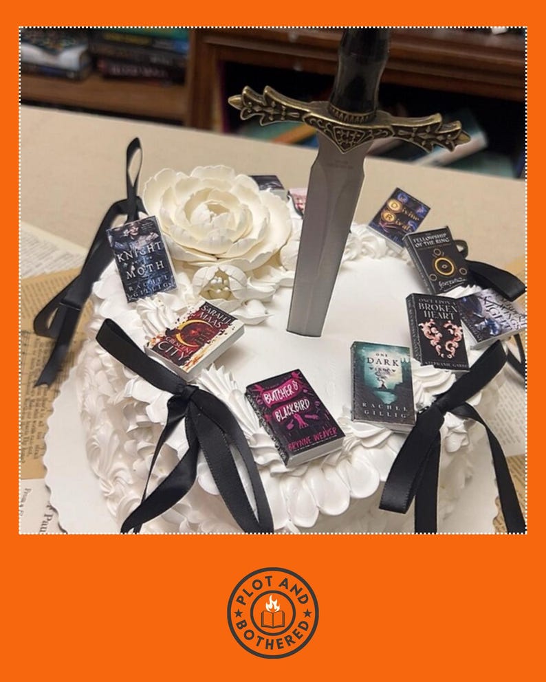 Peut inclure: Un g&acirc;teau blanc d&eacute;cor&eacute; de couvertures de livres miniatures, de rubans noirs et d'une &eacute;p&eacute;e d&eacute;corative. Le g&acirc;teau est orn&eacute; d'une grande fleur blanche. Les livres miniatures ont des titres comme "Knight of Moth" et "City".