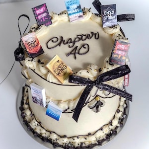 Peut inclure: Un g&acirc;teau rond &agrave; deux niveaux avec un gla&ccedil;age blanc, d&eacute;cor&eacute; d'accents noirs et de petits livres. Le g&acirc;teau porte l'inscription "Chapter 40" et est orn&eacute; de rubans noirs.