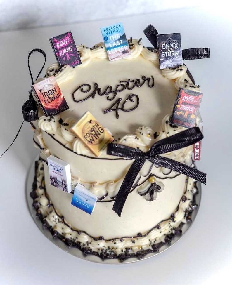 Peut inclure: Un g&acirc;teau rond &agrave; deux niveaux avec un gla&ccedil;age blanc, d&eacute;cor&eacute; d'accents noirs et de petits livres. Le g&acirc;teau porte l'inscription "Chapter 40" et est orn&eacute; de rubans noirs.