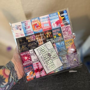 Peut inclure: Un sac en plastique transparent rempli de couvertures de livres miniatures, avec des titres tels que "Confess", "Heart Bones" et "It Ends With Us". Une carte avec "Plot Bothered Book Merch Shop" est visible. Les livres sont de Colleen Hoover.