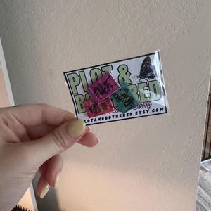 May include: A clear plastic package containing three small rectangular stickers. The stickers are pink, green, and teal, with text that reads "Wings and Ruin", "Court of Fury", and "Mist of Roses". The package also has a logo that says "Plot & Bothered Shop" and the website address.