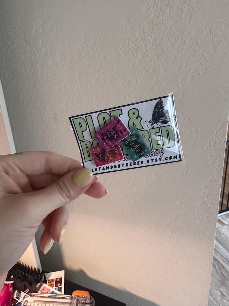 May include: A clear plastic package containing three small rectangular stickers. The stickers are pink, green, and teal, with text that reads "Wings and Ruin", "Court of Fury", and "Mist of Roses". The package also has a logo that says "Plot & Bothered Shop" and the website address.