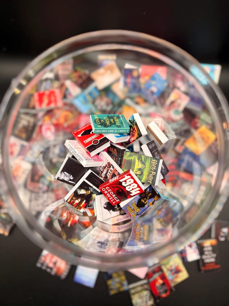 Peut inclure: Un bol en verre transparent rempli de r&eacute;pliques de livres miniatures. Les livres ont des couvertures color&eacute;es et des titres tels que "1984" de George Orwell, "Fahrenheit 451" et "A Court of Mist and Fury". Les livres sont &eacute;parpill&eacute;s dans le bol.