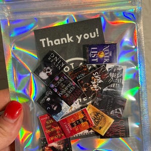 Peut inclure: Un sachet transparent iris&eacute; contenant des r&eacute;pliques de livres miniatures et une carte de remerciement. Les minuscules livres pr&eacute;sentent des titres tels que "A Court of Thorns and Roses" et "Tower of Dawn". La carte dit "Thank you!"