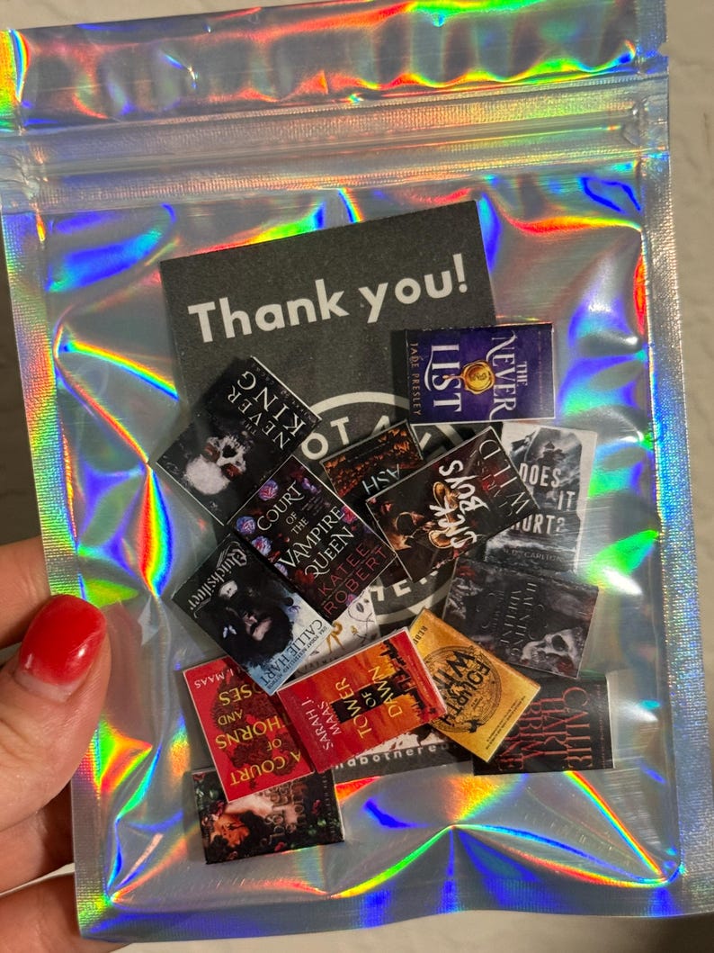 Peut inclure: Un sachet transparent iris&eacute; contenant des r&eacute;pliques de livres miniatures et une carte de remerciement. Les minuscules livres pr&eacute;sentent des titres tels que "A Court of Thorns and Roses" et "Tower of Dawn". La carte dit "Thank you!"