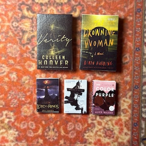 May include: Miniature book collection featuring titles like "Verity" by Colleen Hoover, "Drowning Woman" by Robyn Harding, "The Lord of the Rings" by J.R.R. Tolkien, "The Color Purple" by Alice Walker, and another book. The books are arranged on a patterned orange and brown surface.