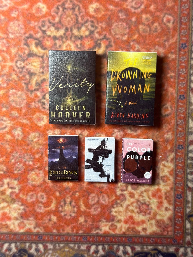 May include: Miniature book collection featuring titles like "Verity" by Colleen Hoover, "Drowning Woman" by Robyn Harding, "The Lord of the Rings" by J.R.R. Tolkien, "The Color Purple" by Alice Walker, and another book. The books are arranged on a patterned orange and brown surface.