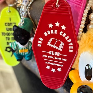 Op de afbeelding: Een verzameling sleutelhangers en bedels. Een rode sleutelhanger met de tekst "ONE MORE CHAPTER CLUB" en een boekillustratie staat centraal. Andere sleutelhangers zijn een gele met tekst en een zwart kattenfiguur.