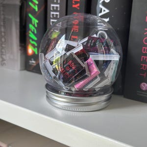 Peut inclure: Un contenant sphérique transparent rempli de petits objets en forme de livre colorés, posé sur un couvercle argenté. Le contenant est placé sur une étagère blanche devant une rangée de livres avec des titres tels que "LIGHT FEAST" et "IT FEEL".