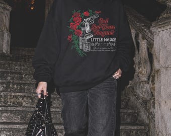 May include: A black crewneck sweatshirt featuring a graphic print of red roses, a handgun, a bottle, a glass, and an eye. The text reads Cat Got Your Tongue? LITTLE MOUSE, along with smaller script text. The sweatshirt is paired with black jeans.