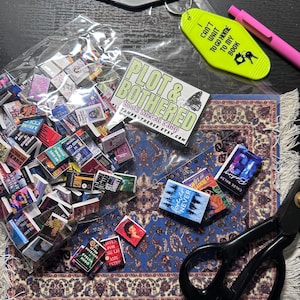 May include: A collection of miniature books, keychains, and a pen. The keychains read "Chronic Reader" and "I can't wait to go home to my book." A "Plot & Bothered" shop sticker is also present. The books are scattered on a blue rug.