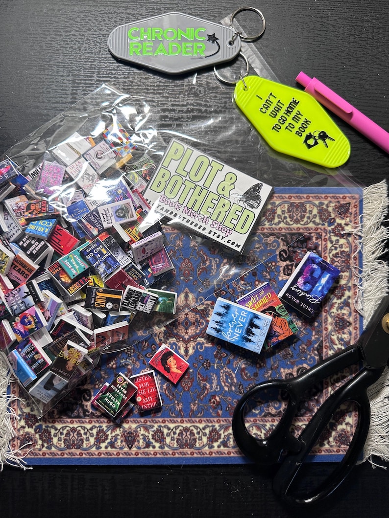 May include: A collection of miniature books, keychains, and a pen. The keychains read "Chronic Reader" and "I can't wait to go home to my book." A "Plot & Bothered" shop sticker is also present. The books are scattered on a blue rug.
