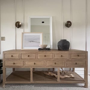 May include: A light-colored wooden console table with multiple drawers and a lower shelf. The table is adorned with a framed picture, a woven bowl, a dark vase, and a book. Two sconces are mounted on the wall above the table.