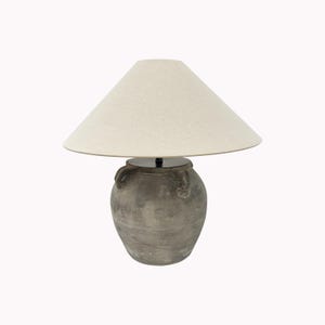 May include: A gray ceramic table lamp with a cream-colored lampshade. The lamp base has a textured, aged appearance with two small handles. The lampshade is conical and made of a light-colored fabric. The lamp is designed for indoor use.