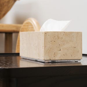 May include: A beige travertine tissue box holder with a white tissue box inside. The holder is sitting on a dark wood surface.