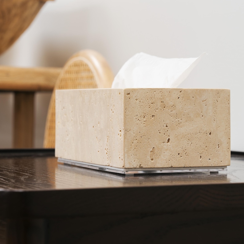 May include: A beige travertine tissue box holder with a white tissue box inside. The holder is sitting on a dark wood surface.