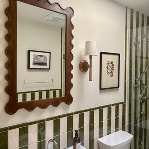 May include: A bathroom with a large, ornate, brown-framed mirror. The walls feature green and white striped tiles and a framed botanical print. A gold sconce with a cream shade and a white sink with chrome fixtures are also visible.