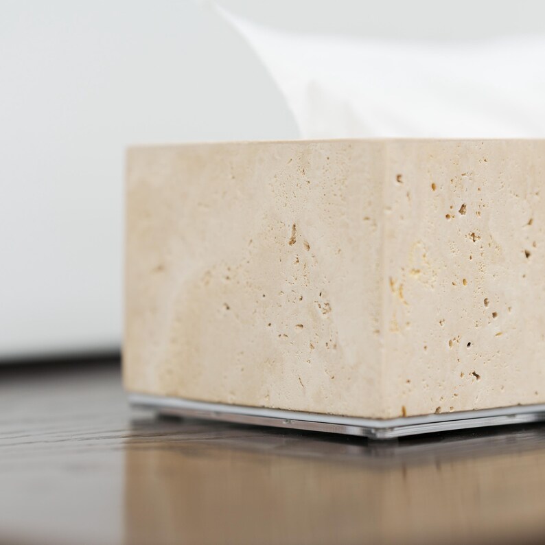 May include: A beige travertine tissue box holder with a clear acrylic base. The holder has a rough, textured surface.
