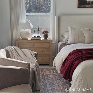 May include: A bedroom with a white headboard, a white bedspread, and a red throw blanket. A wooden dresser with a white lamp sits next to the bed. The room has a window with a view of trees.