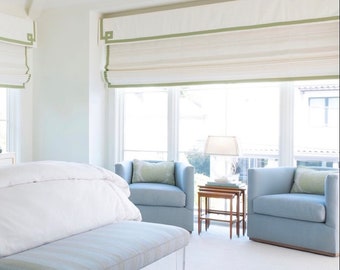 May include: A bedroom with a white bed, a light blue bench, two light blue armchairs, and a wooden end table. The windows have white roman shades with a green trim.