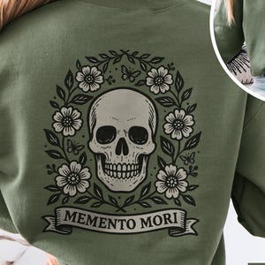May include: Military green sweatshirt featuring a skull design. The back of the sweatshirt displays a skull surrounded by flowers and butterflies, with the words "Memento Mori" on a banner. A smaller skull design is on the front. The sweatshirt is a solid color.