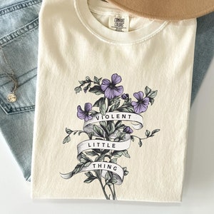 May include: A cream colored t-shirt with a floral design and the text "VIOLENT LITTLE THING" on a banner.