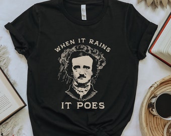 May include: Black t-shirt featuring a portrait of Edgar Allan Poe with the text "WHEN IT RAINS IT POES" in a vintage style. The design is in a cream color. The shirt is displayed on a white surface with books and a coffee cup.