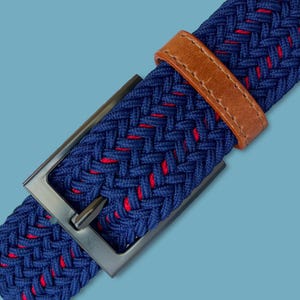 May include: Close-up of a blue woven belt with red accents. The belt features a brown leather loop and a rectangular, gunmetal-colored buckle. The woven design adds texture and visual interest to the accessory.