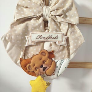 Può includere: Decorazione per la cameretta beige e bianca con un grande fiocco a motivi di stelle. Sotto il fiocco, uno stendardo con il nome "Raffaele". Un orsetto di feltro dorme su un cuscino, con una stella gialla appesa.