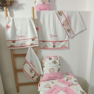 May include: A collection of white fabric items including towels, drawstring bags, and small pouches, all featuring a pink hot air balloon pattern and the name Ginevra embroidered in pink script. The set includes various sizes of rectangular towels.