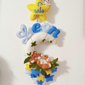 Può includere: Una luna crescente di feltro bianco con un nome "Leon" di feltro blu e giallo e una piccola stella gialla con il testo "E nato 27.9.2024". Un cucciolo di leone di feltro marrone è sdraiato su una foglia verde di feltro sulla luna. Ci sono stelle di feltro blu e giallo intorno alla luna.