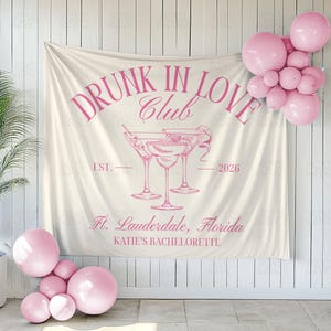 May include: A cream-colored banner with pink text that reads "DRUNK IN LOVE Club" with martini glasses and the text "Ft. Lauderdale, Florida KATIE'S BACHELORETTE". Pink balloons are clustered on the wall and floor.