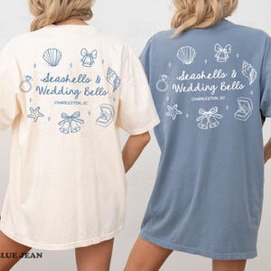 K&ouml;nnte beinhalten: Zwei &uuml;bergro&szlig;e T-Shirts, eines in Elfenbein und eines in Blau, mit dem Text "Seashells & Wedding Bells" und zugeh&ouml;rigen Grafiken. Das Design umfasst Muscheln, Glocken, einen Ring und einen Seestern. Die Shirts sind in Gr&ouml;&szlig;e XL.