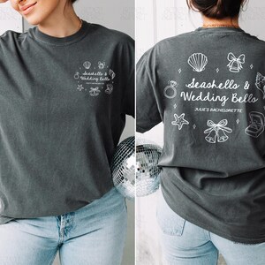 K&ouml;nnte beinhalten: Dunkelgraues T-Shirt mit dem Text "Seashells & Wedding Bells" und zugeh&ouml;rigen Grafiken. Auf der R&uuml;ckseite des Shirts steht au&szlig;erdem der Text "Julie's Bachelorette". Das Shirt wird mit hellblauen Jeans kombiniert.
