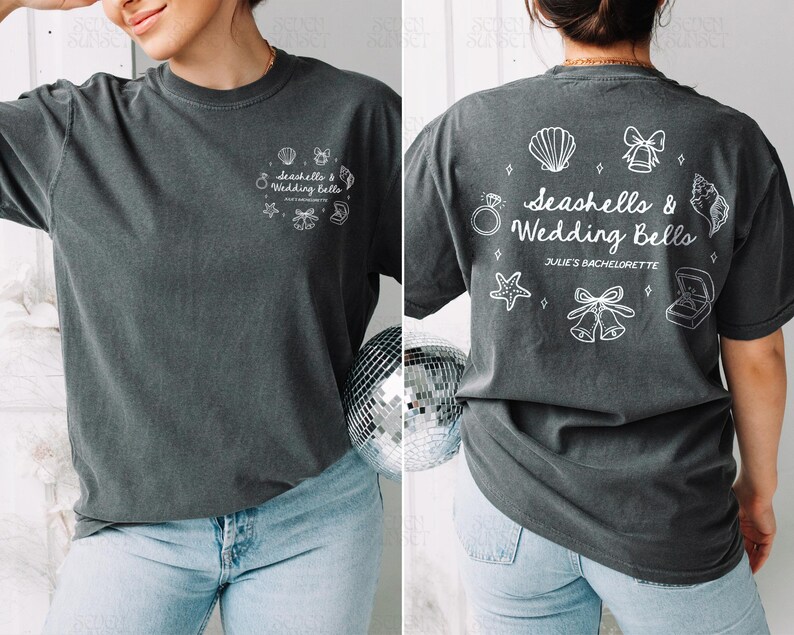 K&ouml;nnte beinhalten: Dunkelgraues T-Shirt mit dem Text "Seashells & Wedding Bells" und zugeh&ouml;rigen Grafiken. Auf der R&uuml;ckseite des Shirts steht au&szlig;erdem der Text "Julie's Bachelorette". Das Shirt wird mit hellblauen Jeans kombiniert.
