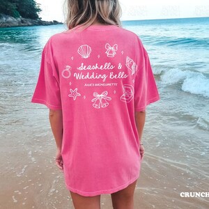 K&ouml;nnte beinhalten: Ein pinkfarbenes T-Shirt mit dem Text "Seashells & Wedding Bells" und wei&szlig;en grafischen Symbolen. Das T-Shirt, Gr&ouml;&szlig;e L, wird an einem Strand getragen. Die Farbe des Shirts ist "Crunchberry".